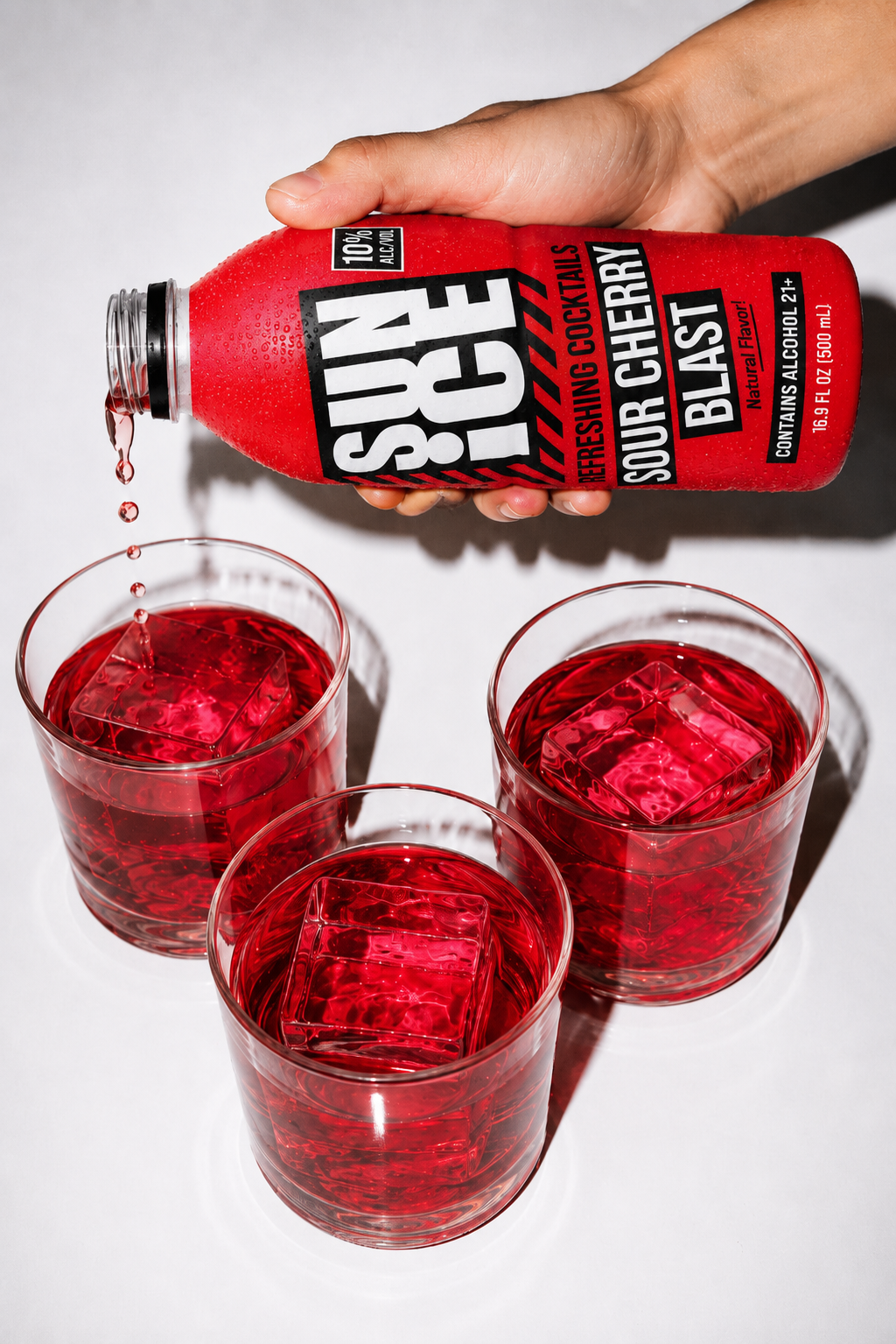 A hand pours Sour Cherry Blast SUN ICE from the bottle into a glass over ice, with two more filled glasses alongside.